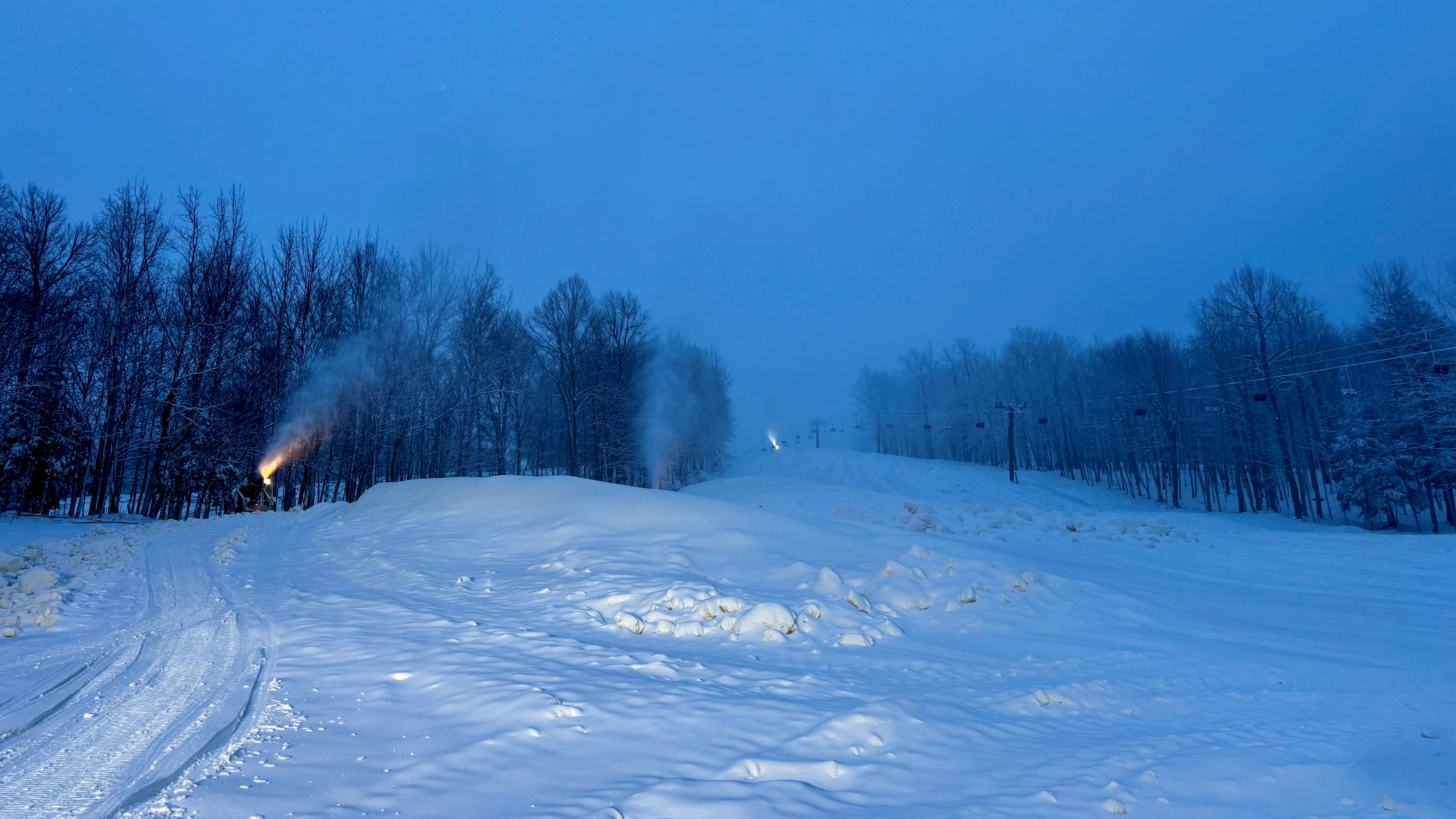 View up a ski run with snowguns running, creating piles of snow.