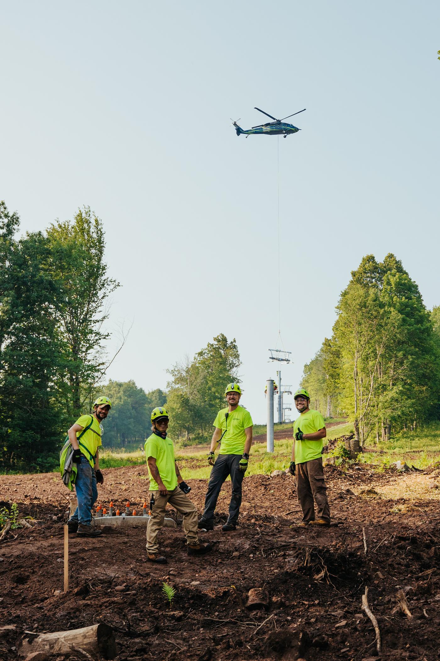 Installation crewmembers stand in foreground while helicopter places tower in background.