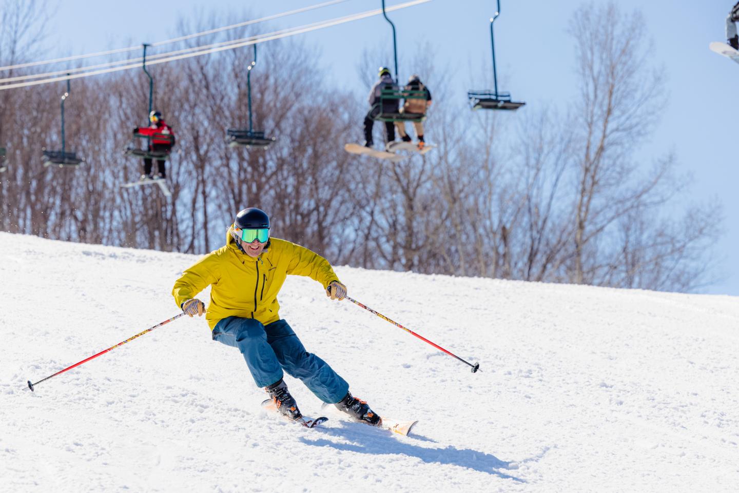 man in yellow goat and blue pants skis with a smile in springtime.