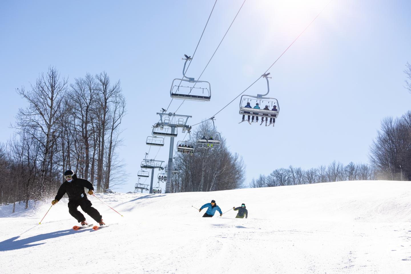 Three skiers speed down the hill under a chairlift in the sun.