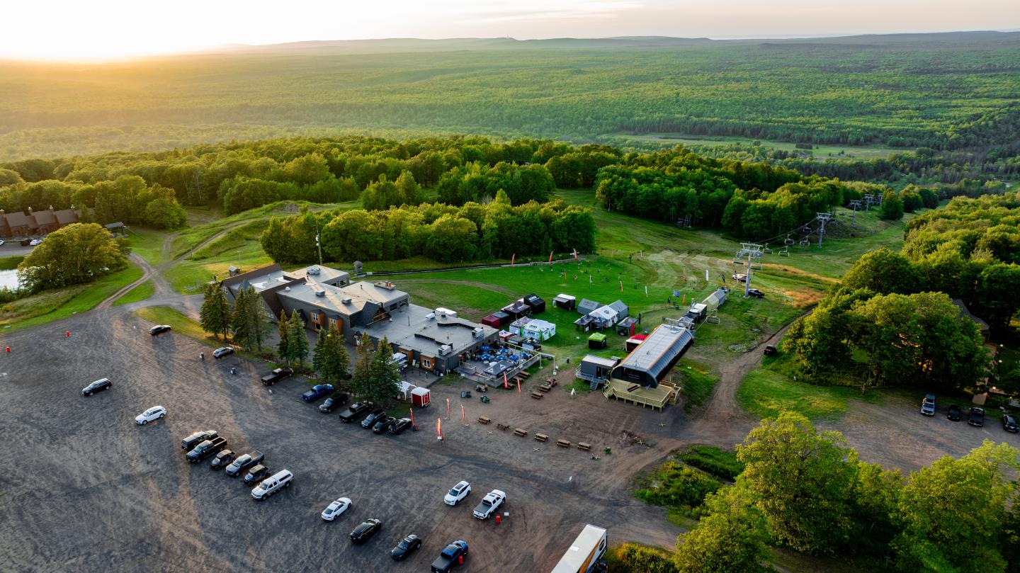 Aerial view of a countryside event with trees and parked cars at sunset.