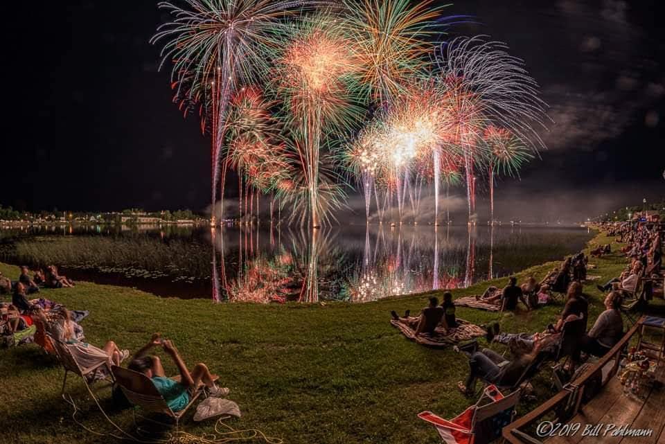 Fireworks lighting up the night sky over a lake, with people watching from grassy banks.