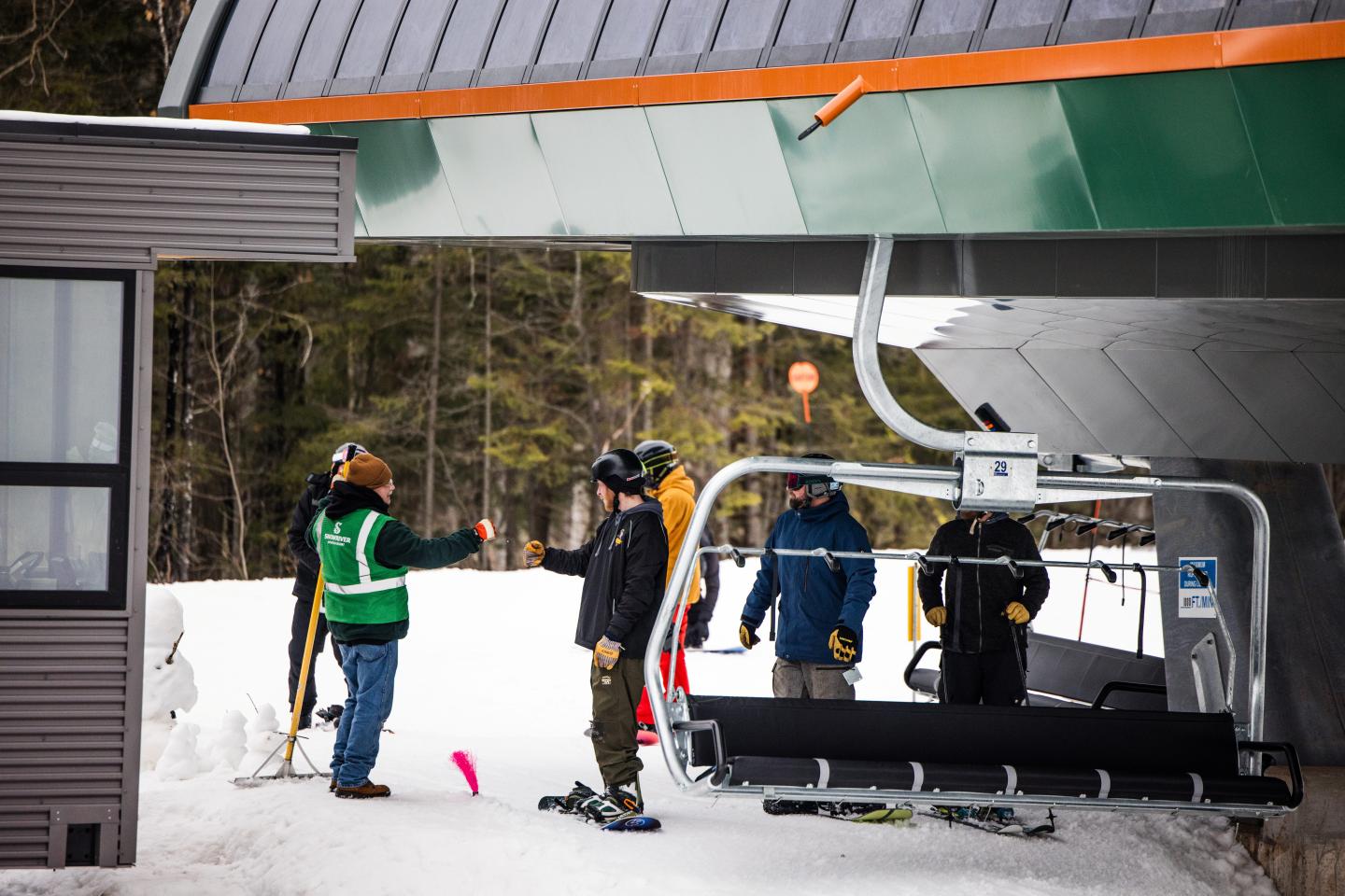 Skiers waiting at a chairlift in a snowy, wooded area.