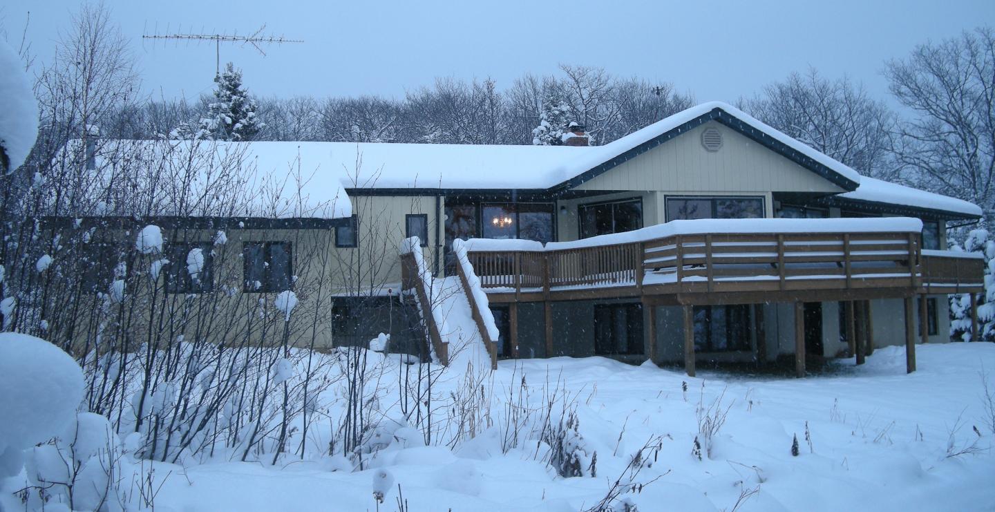 Snow-covered house with a wooden porch surrounded by trees.
