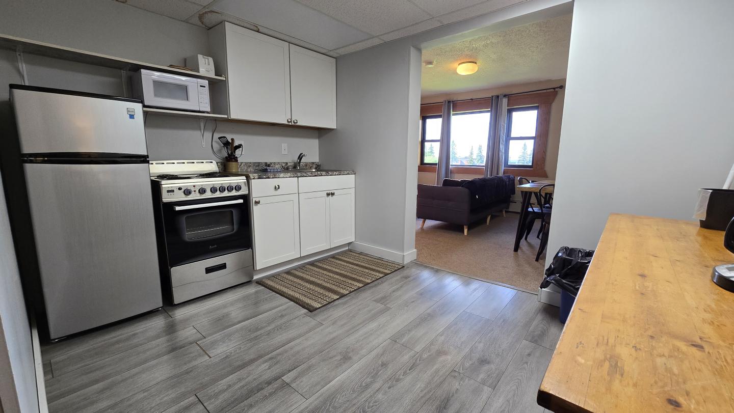 Small kitchen with gray flooring, connected to a living room with large windows.