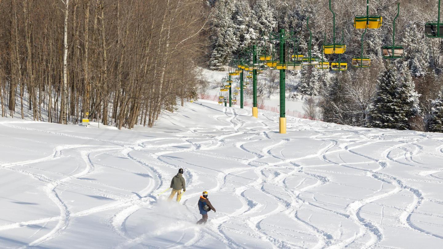 Skiers on snow-covered slope with trees and a lift in the background.