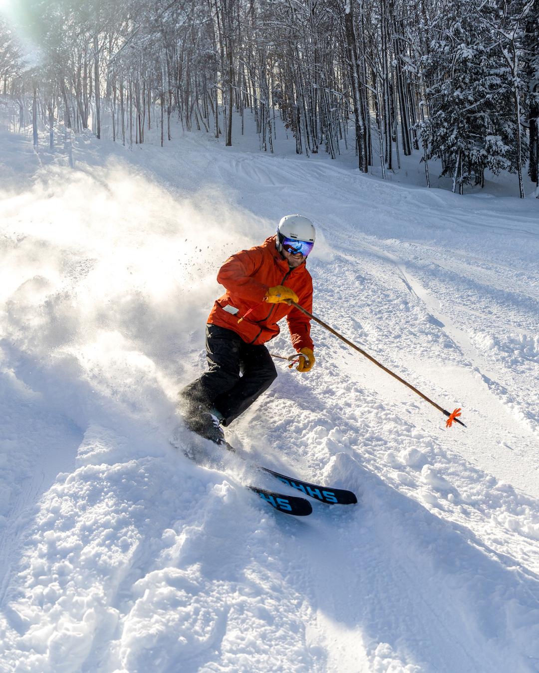 Skier in orange jacket skiing down snowy slope, surrounded by snow-covered trees.