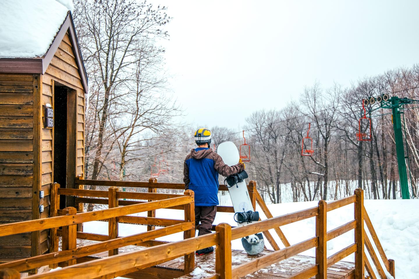 Person holding a snowboard outside a wooden cabin in snowy woods.