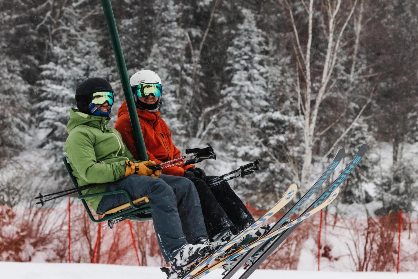Two skiers on a lift, snowy forest background.
