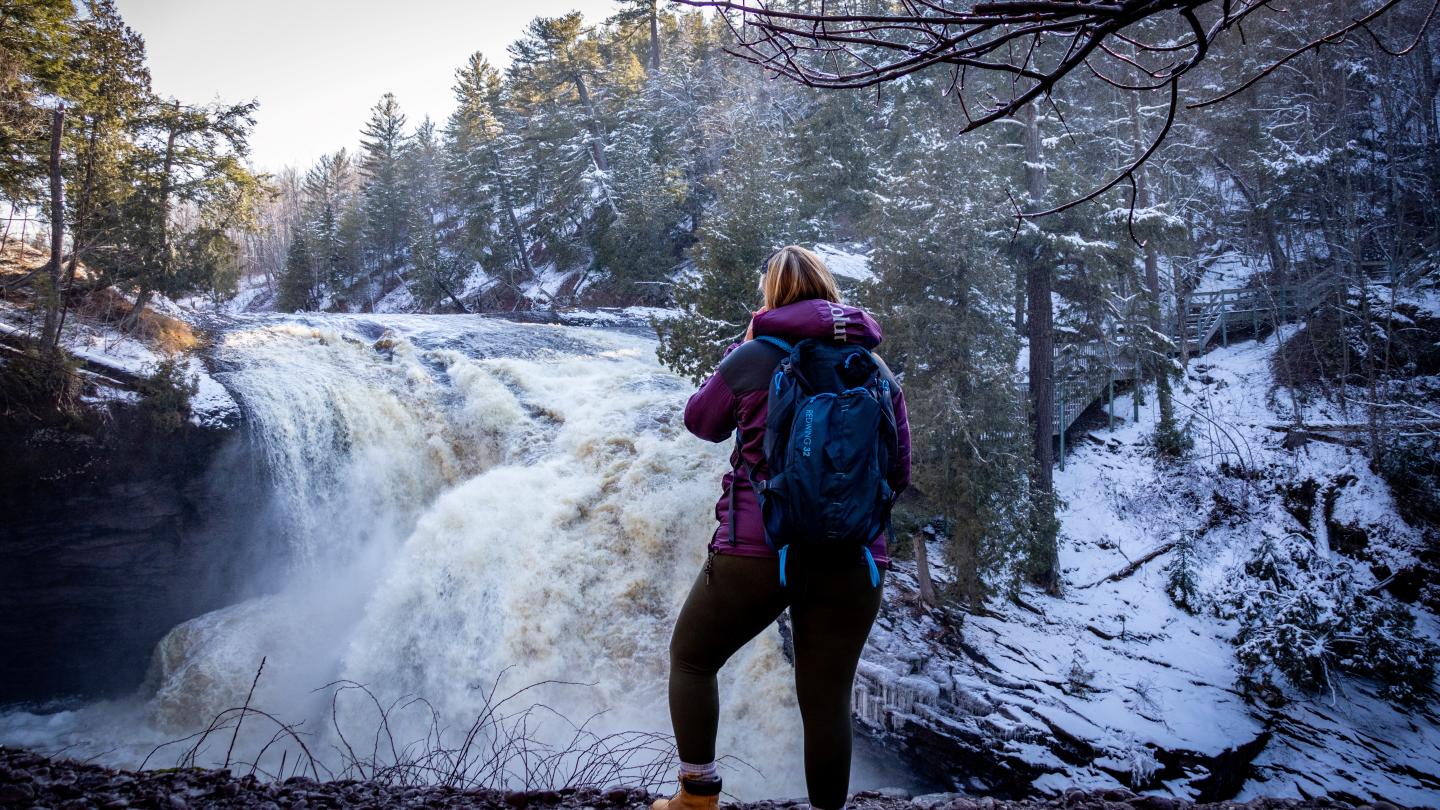 Person with backpack standing near a snow-covered waterfall surrounded by trees.