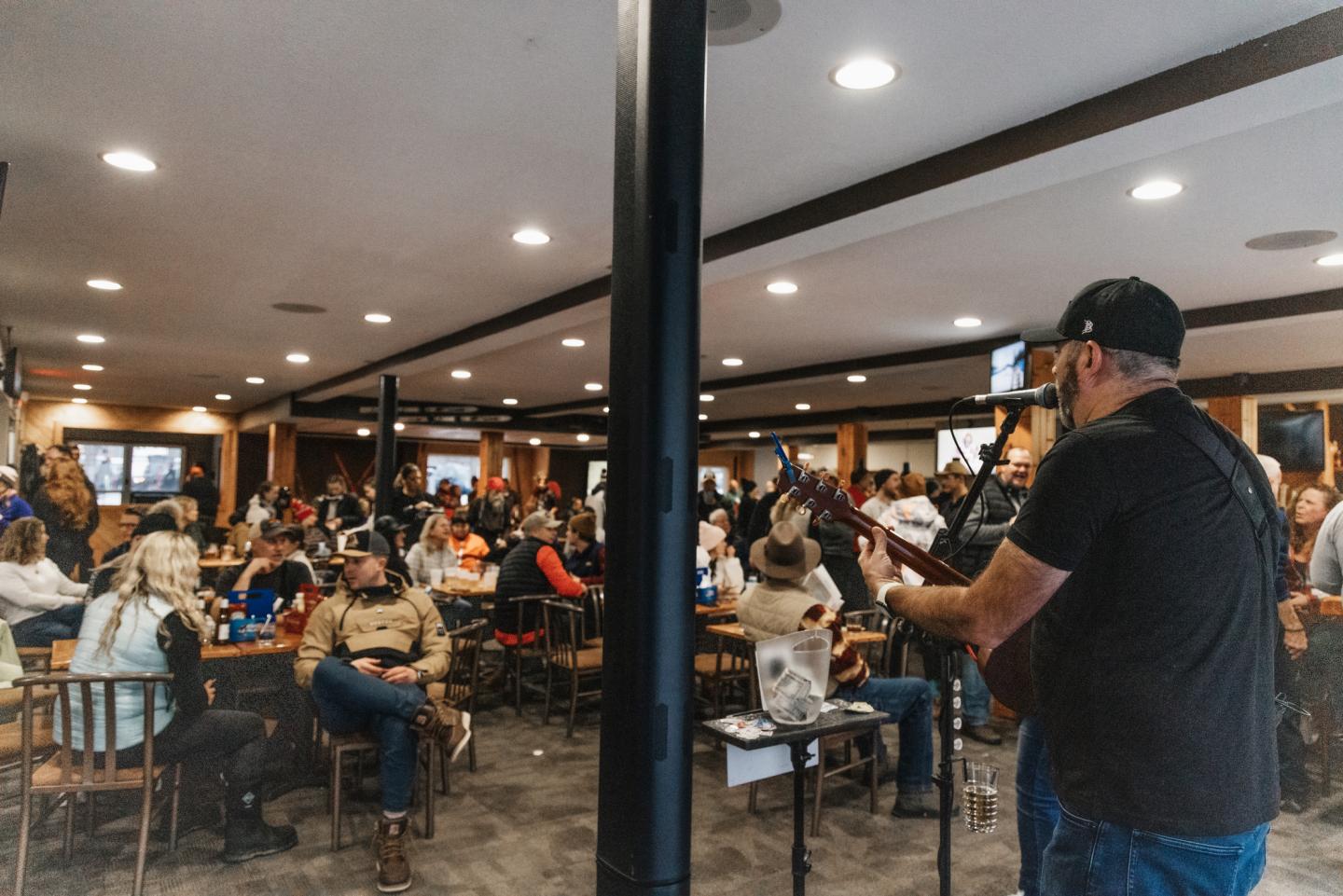 Man playing guitar in a crowded café, people seated at tables listening.