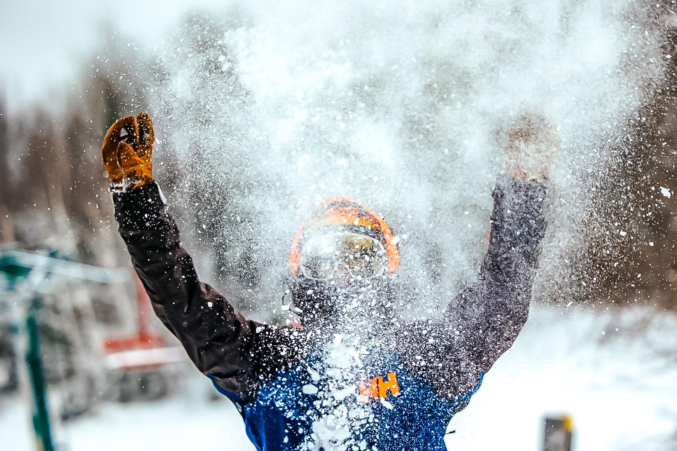 Person throwing snow in the air, wearing winter gear and goggles.