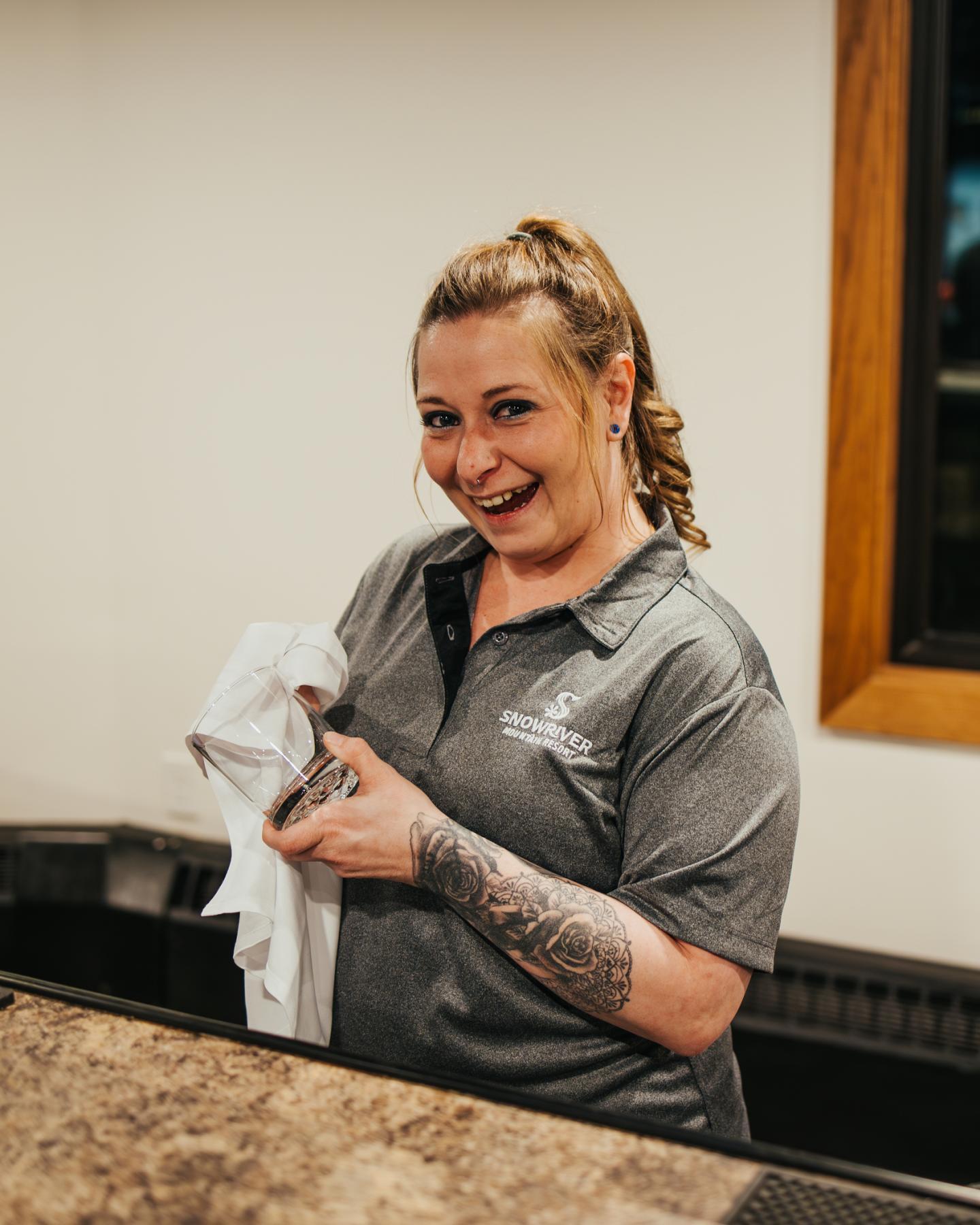 Smiling person behind a counter, drying a glass with a towel.