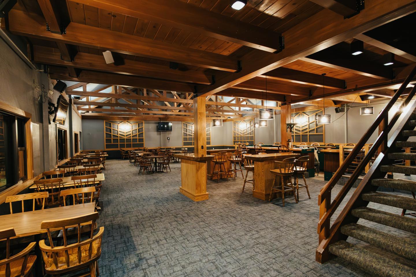 Rustic restaurant interior with wooden beams and tables, carpeted floor, and staircase.