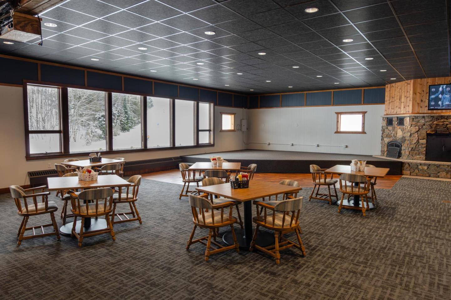 Dining area with wooden tables, chairs, large windows, and a stone fireplace.