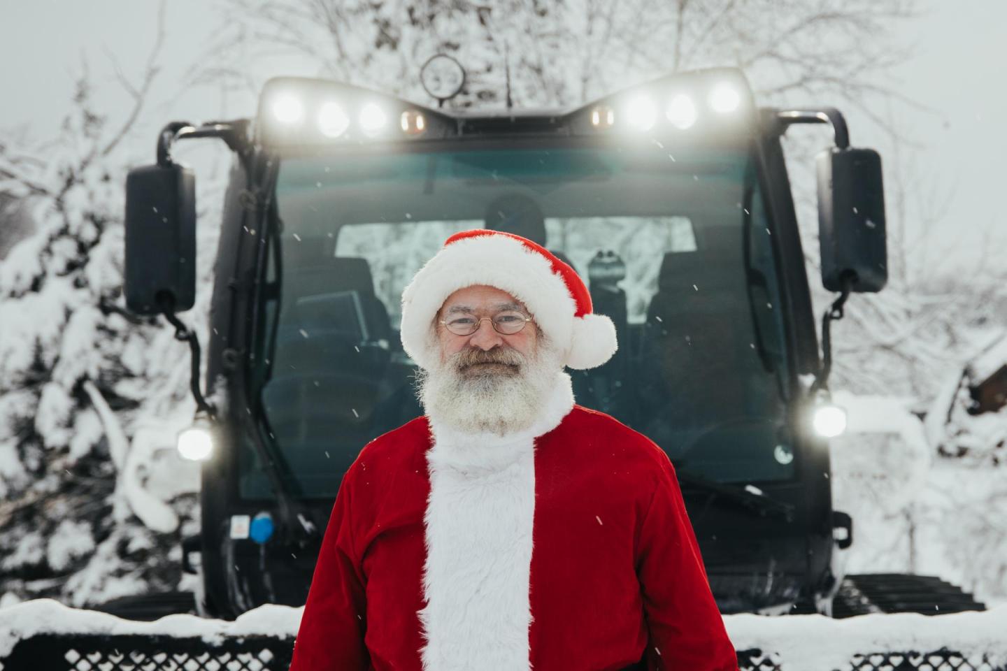 Santa Claus stands in front of a snow-covered tractor.
