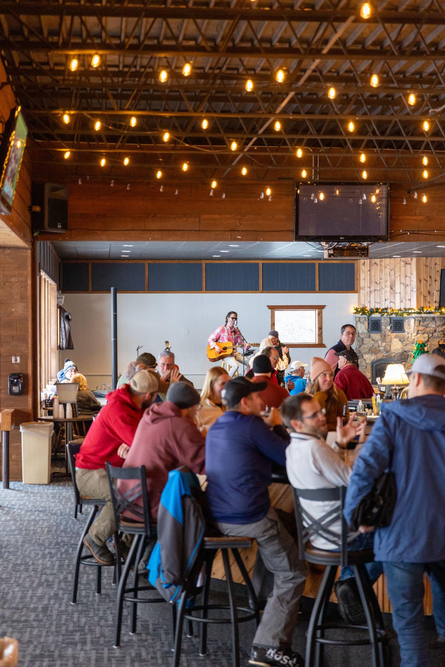 People sit at a bar, watching a musician perform on stage under warm string lights.