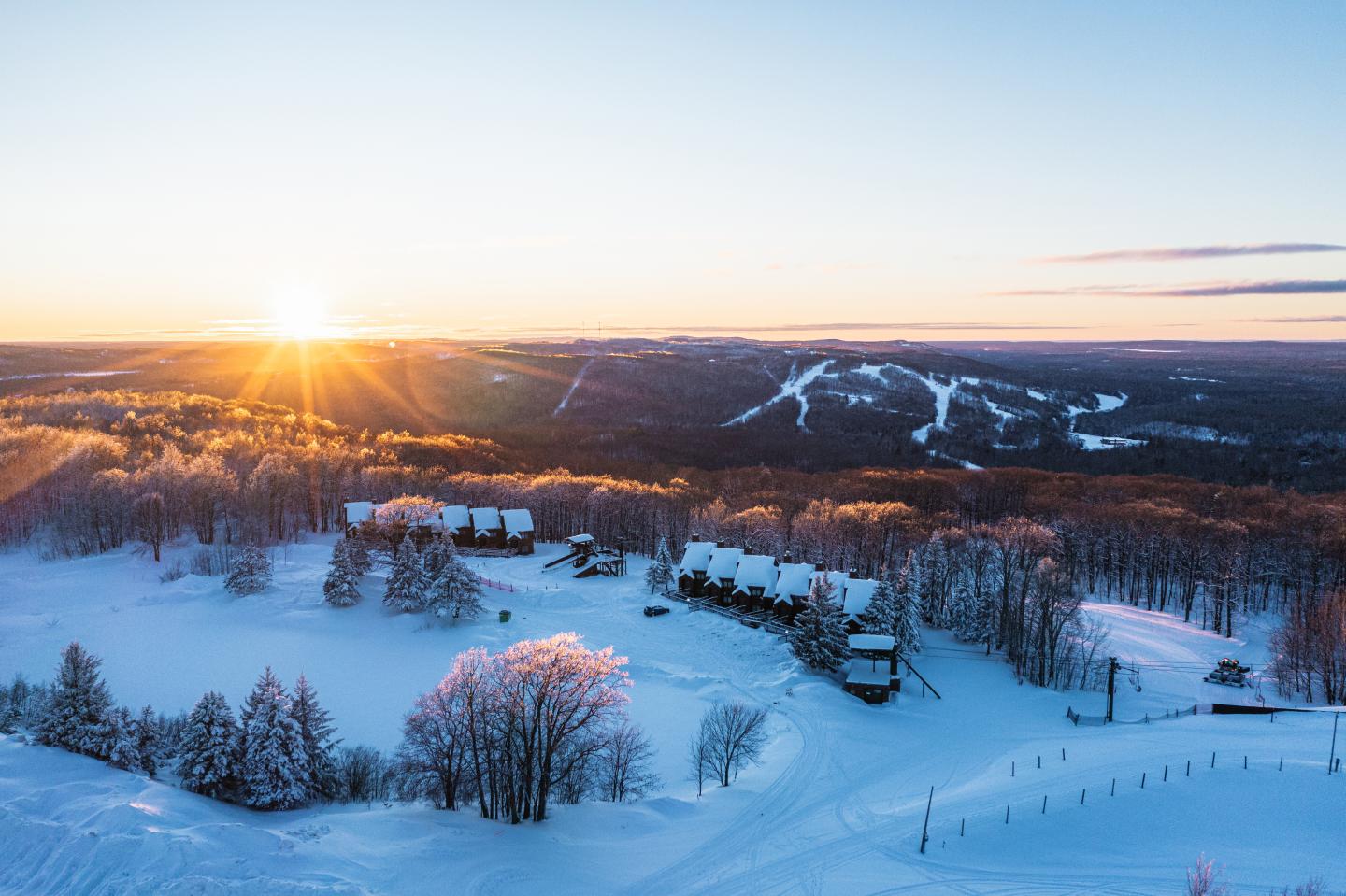 Sunrise over snowy landscape, trees, and cabins with distant hills.