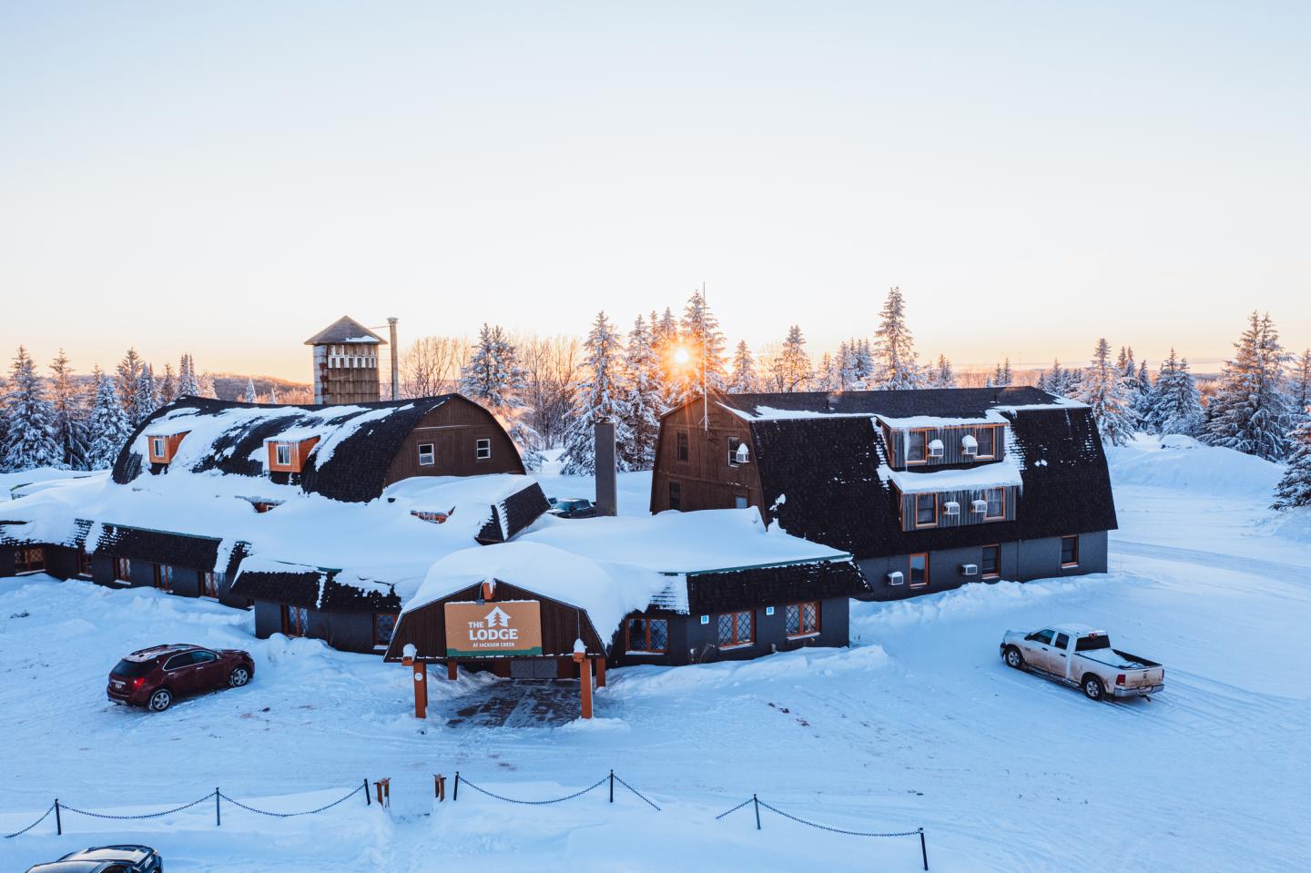 Snow-covered lodge at sunset, surrounded by trees and cars.