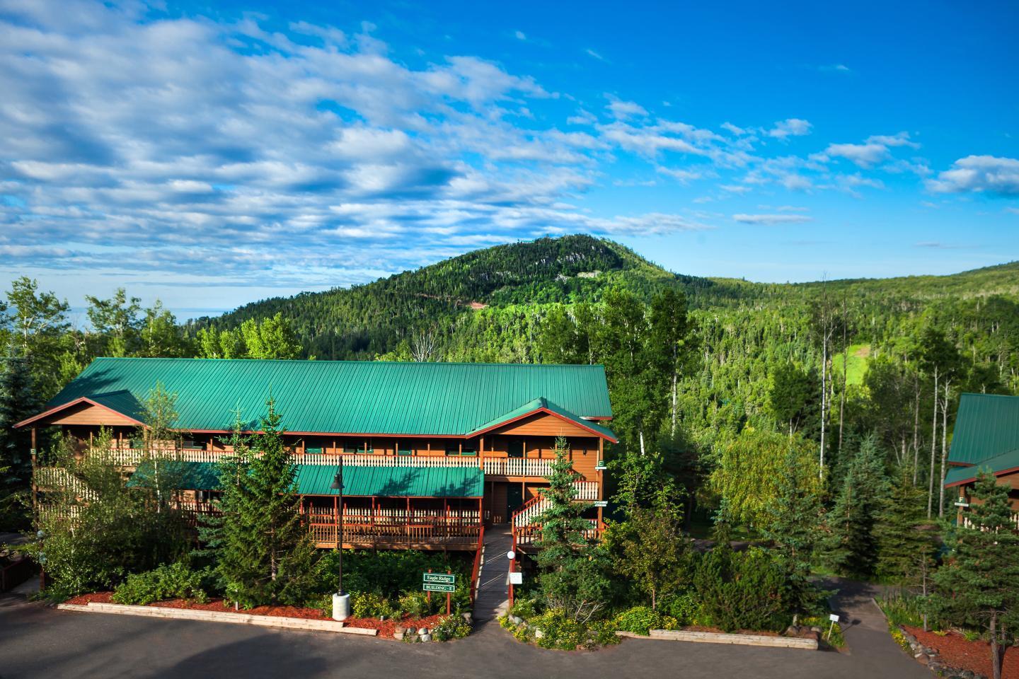 Mountain lodge with green roofs, surrounded by lush forest under a blue sky.