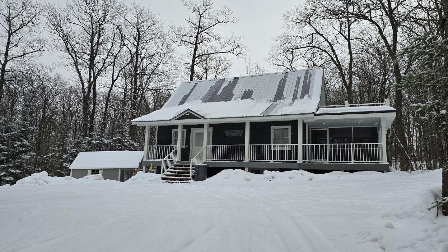 Snow-covered house in a forest, overcast sky, surrounded by bare trees.