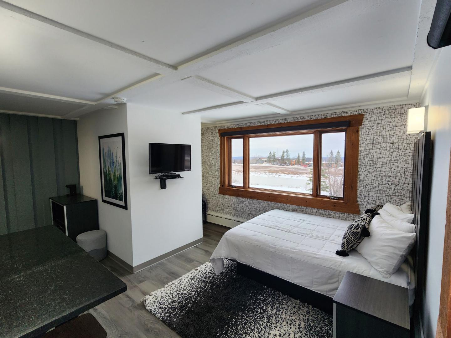 Cozy bedroom with a large window, bed, wall TV, and a small seating area.