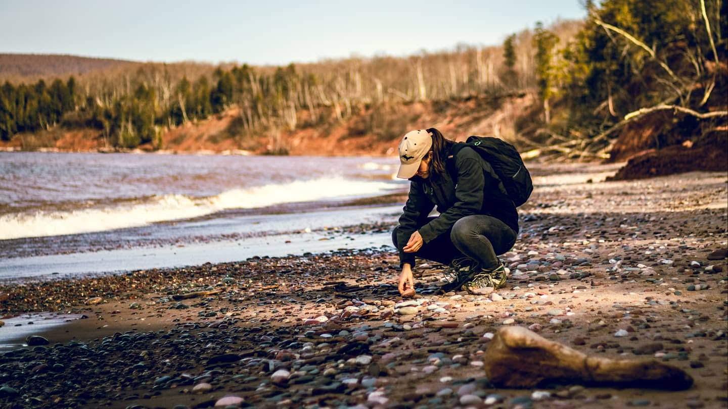 Person kneeling on rocky beach, picking up stones near water.