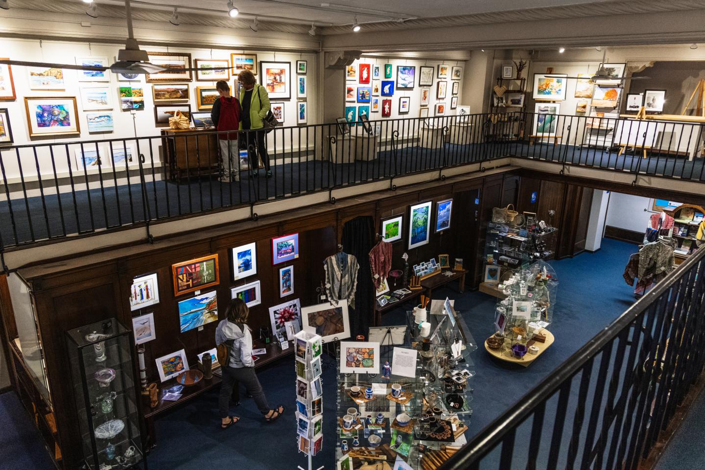 Art gallery interior with colorful framed artworks on walls, two people browsing upstairs.