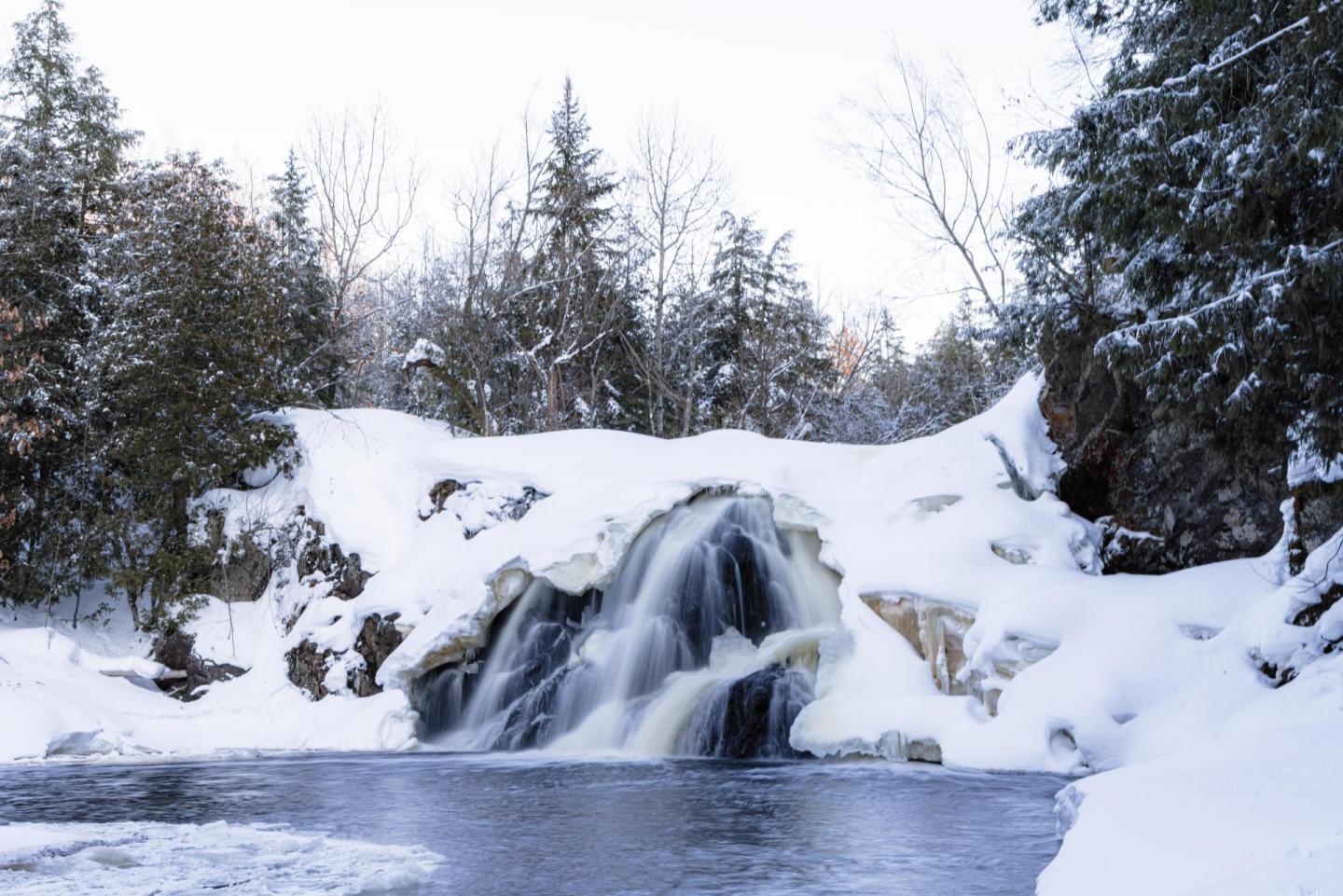 Snow-covered waterfall flowing into a calm river, surrounded by trees.