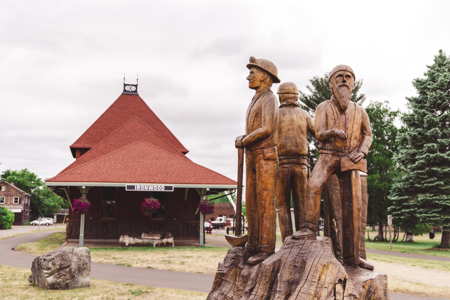 Wooden statue of three figures near a red-roofed building and trees.