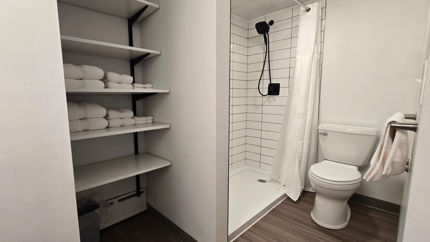 Bathroom with shower, toilet, shelves of towels, and wood-style flooring.