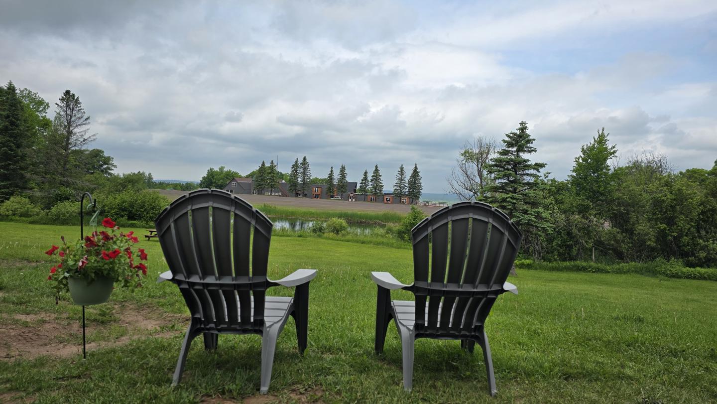 Two black chairs on grass facing a lake and trees under a cloudy sky.