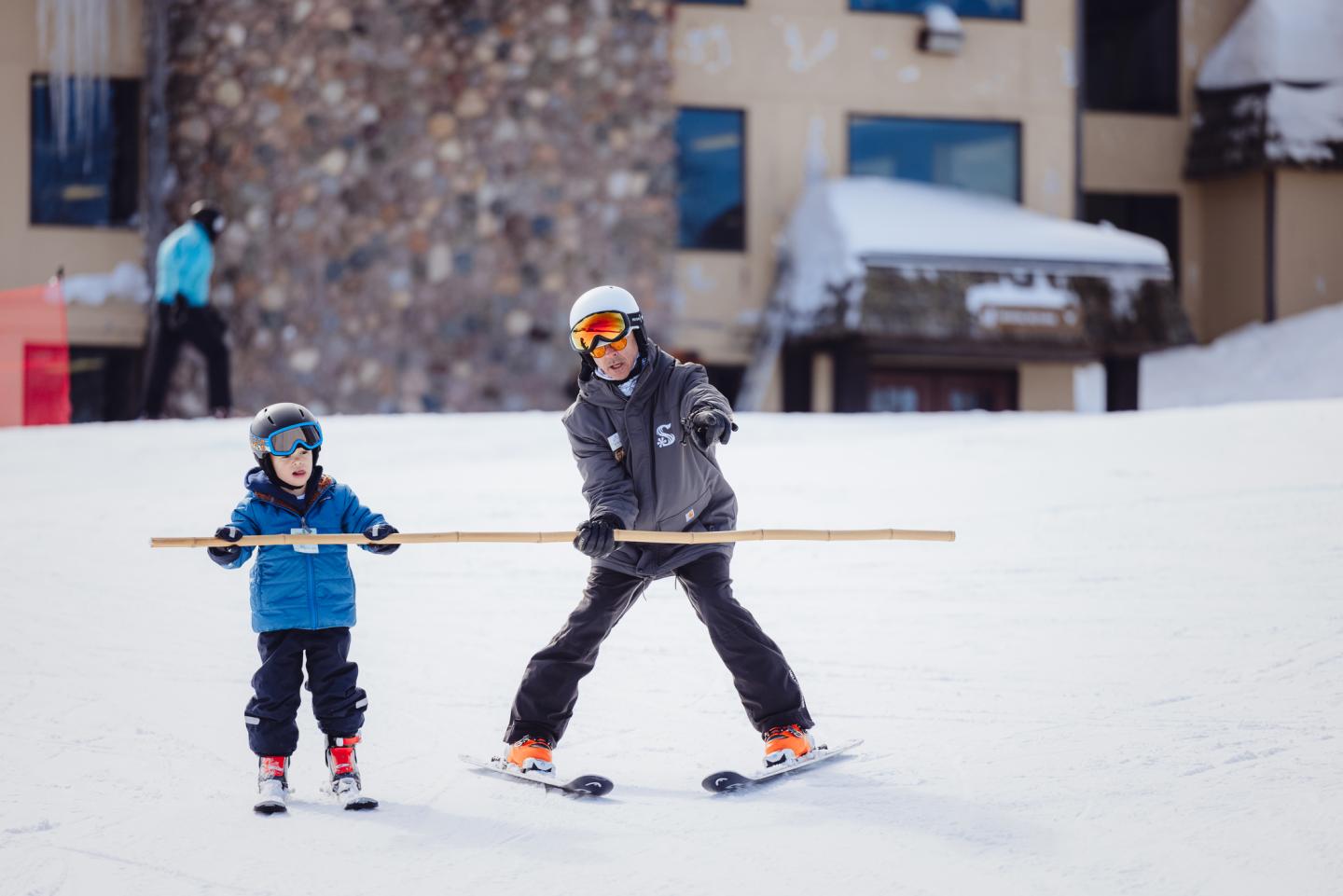 Child and adult skiing on snowy slope with a wooden stick, wearing helmets and goggles.