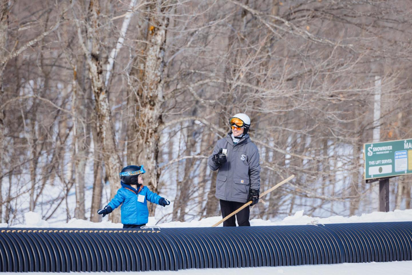 Child and adult skiing on snow against a backdrop of leafless trees.