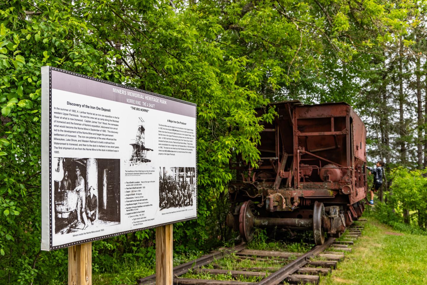 Old train car on tracks near lush green trees and an informational sign.