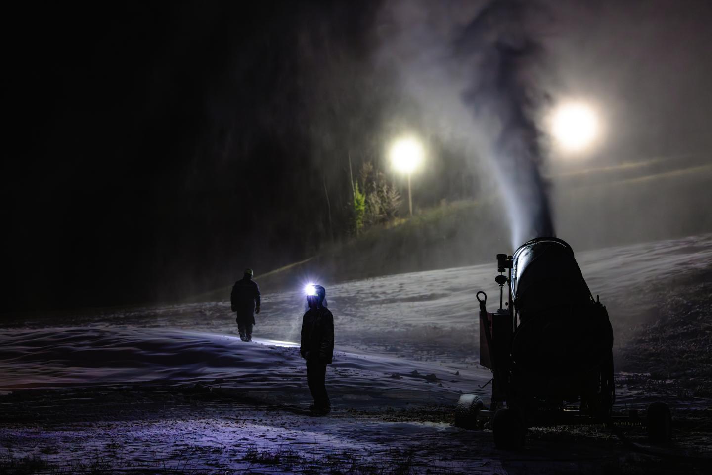Snowgun operating at night, two people with headlamps on a snowy slope.