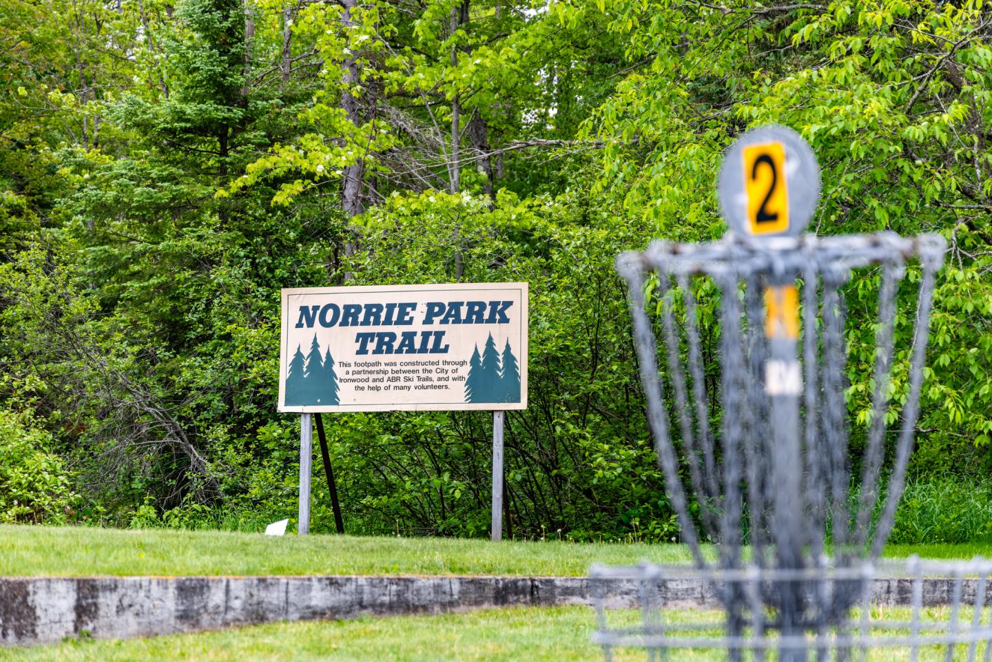 Disc golf basket with the number two, Norrie Park Trail sign, green trees in the background.