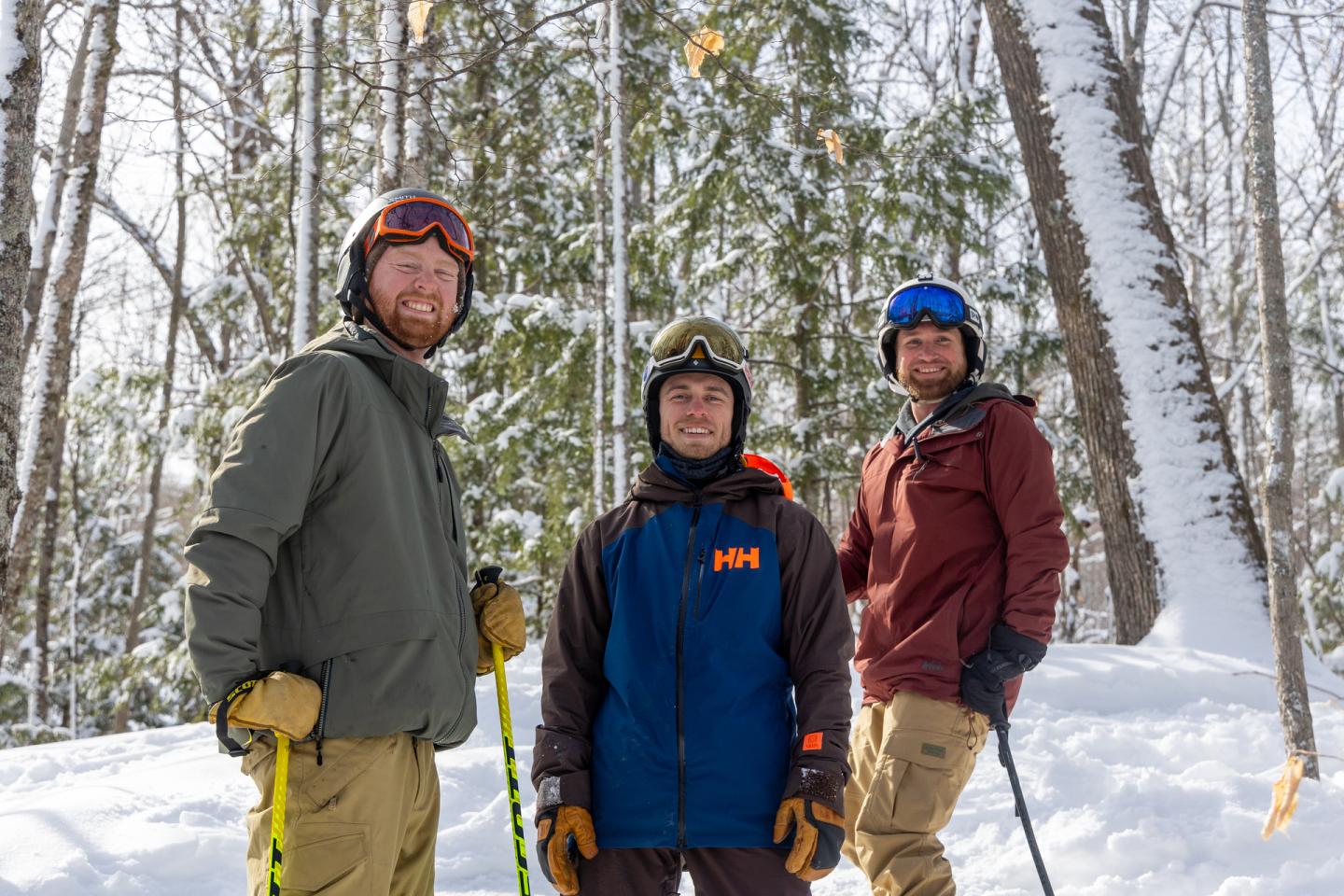 Three skiers standing in snowy woods, wearing winter gear, smiling.