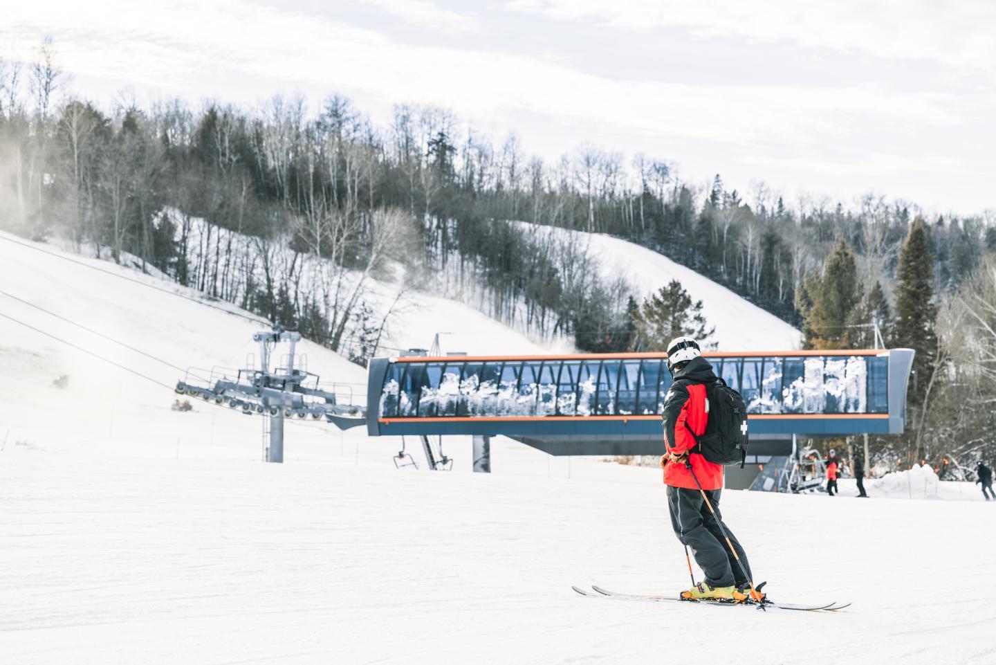 Skier in red jacket on snowy slope, trees and ski lift in background.