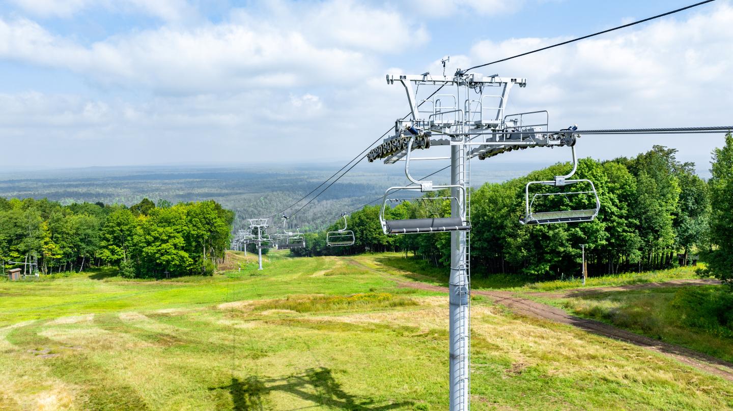 Ski lift over grassy hillside with blue sky and distant trees.