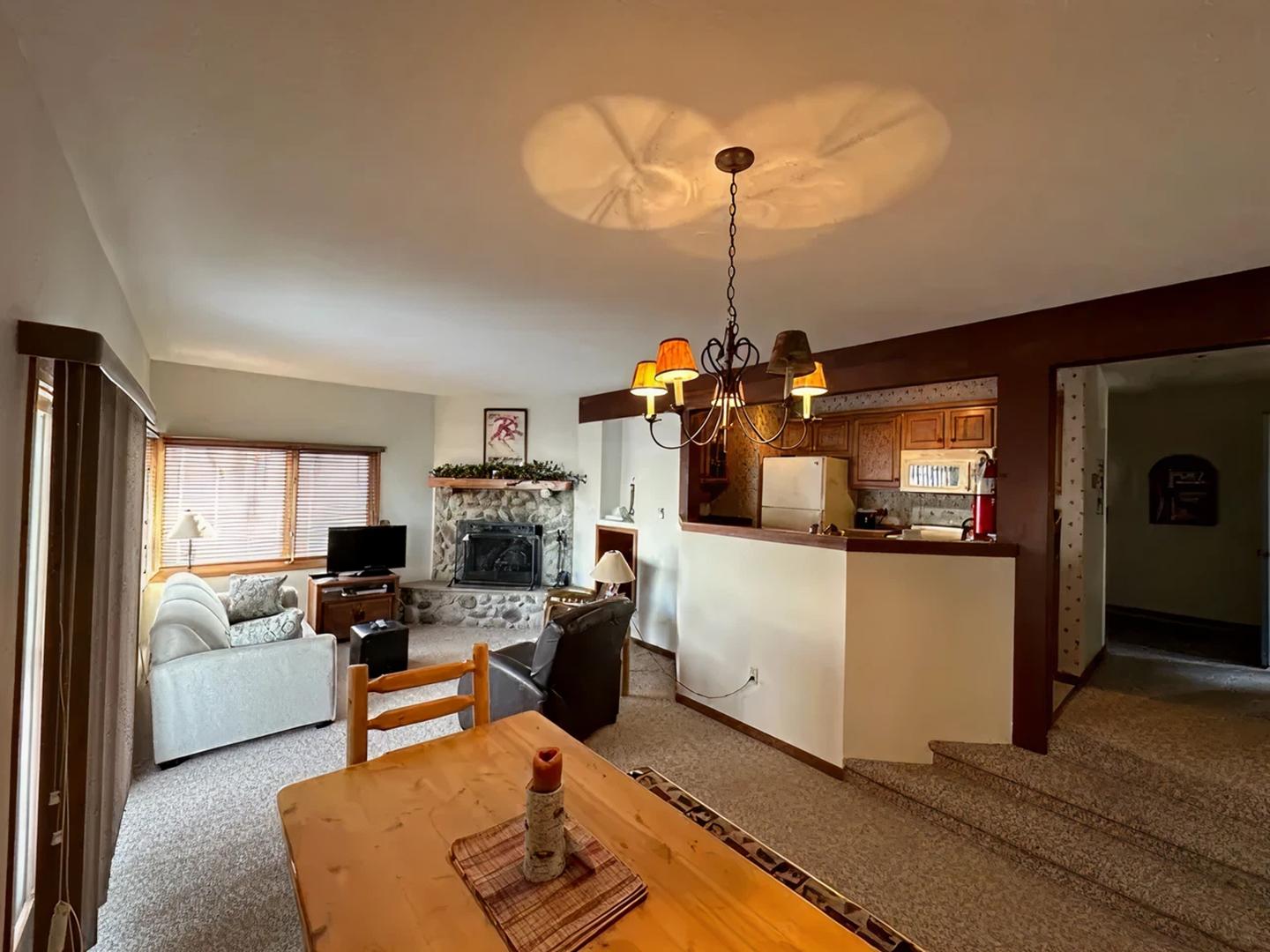 Cozy living room with fireplace, sofa, and dining table. Warm lighting, kitchen view.