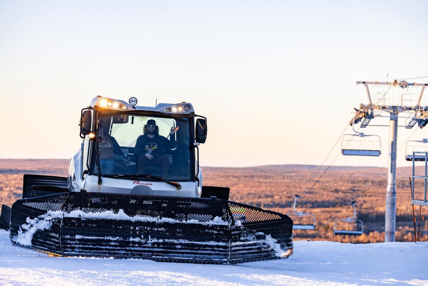 Snowplow grooming a snowy slope at sunset, ski lift in the background.