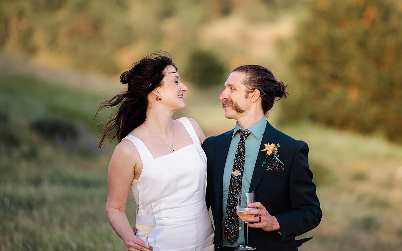 Couple smiling outdoors in formal attire, holding drinks, lush greenery in background.