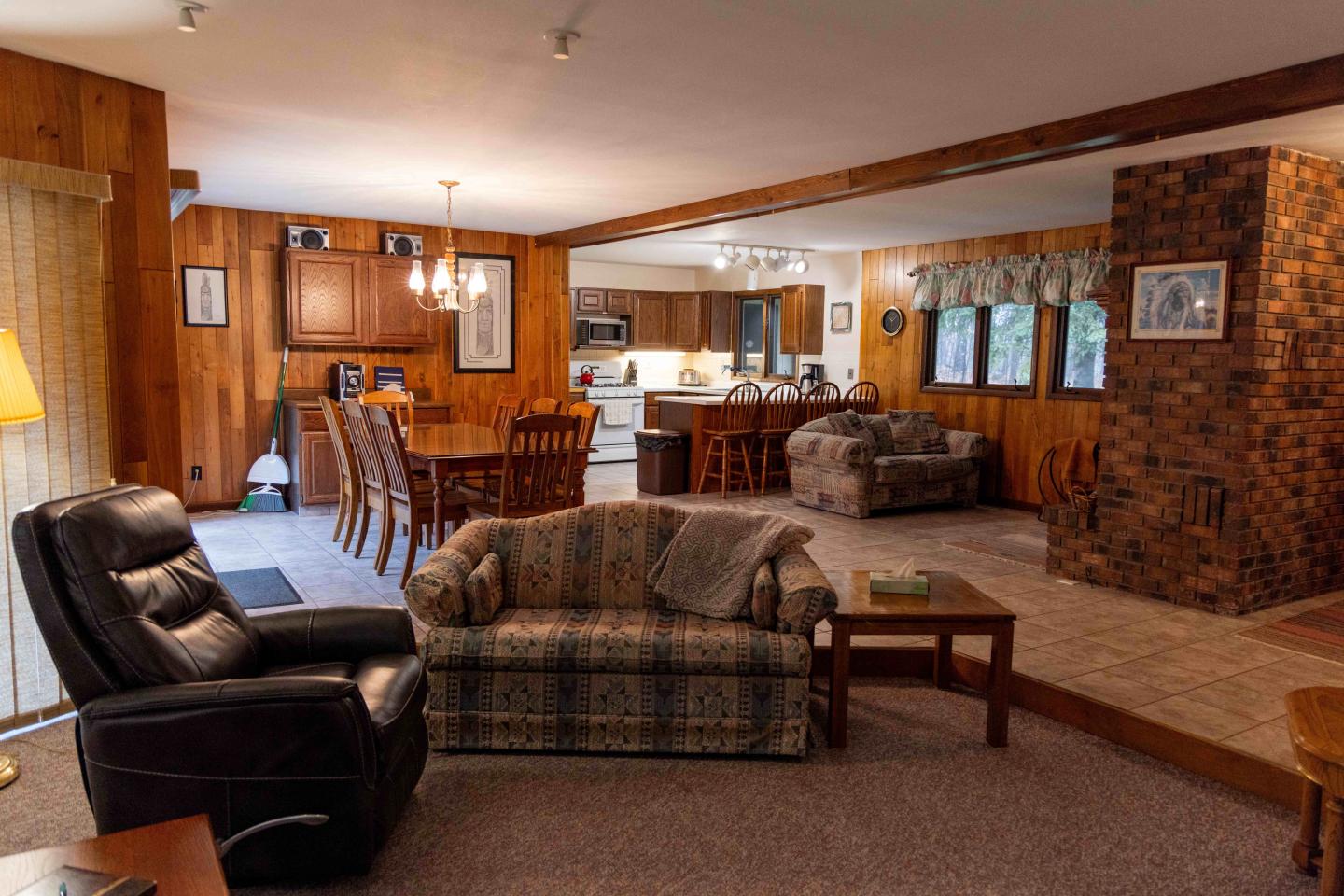 Cozy living room with sofas, dining table, and open kitchen. Wood-paneled walls and brick accents.