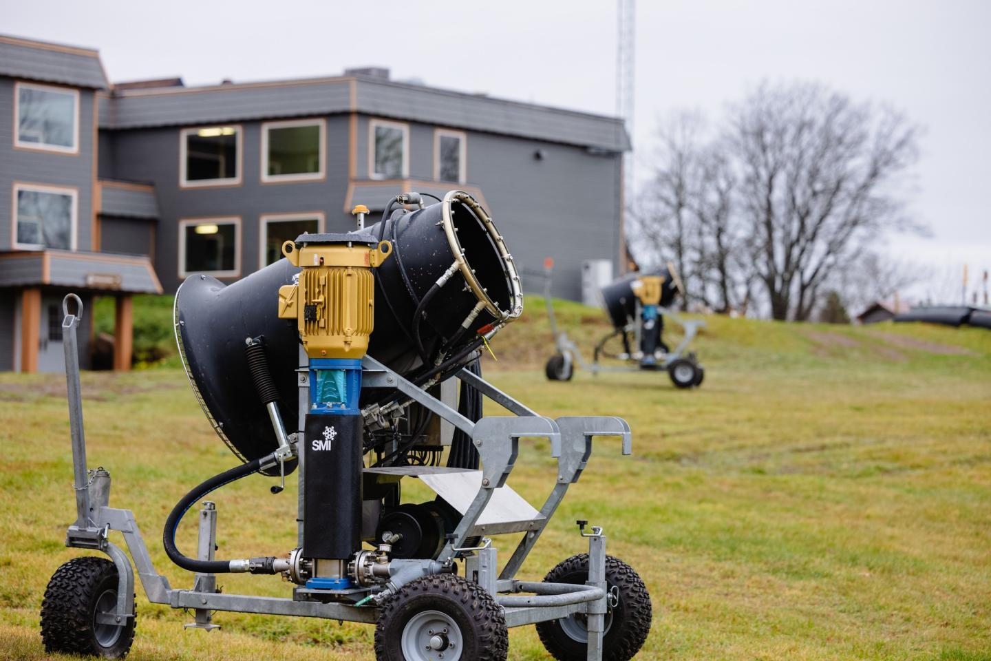 Snow machines on grass with buildings and trees in the background.