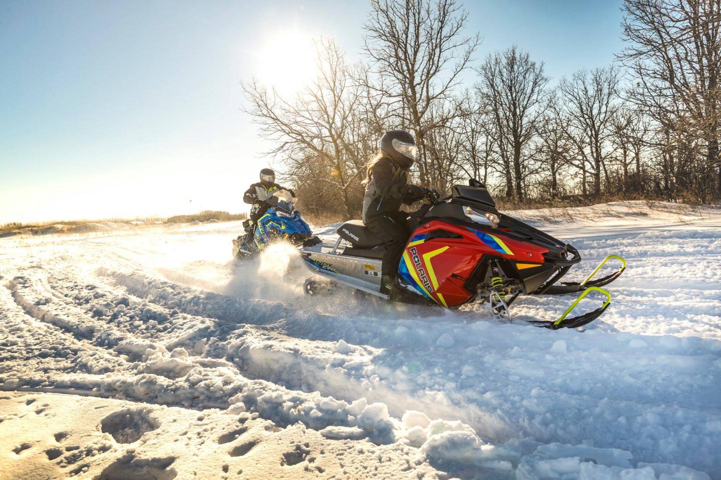 Two people riding snowmobiles on a sunny, snowy landscape.