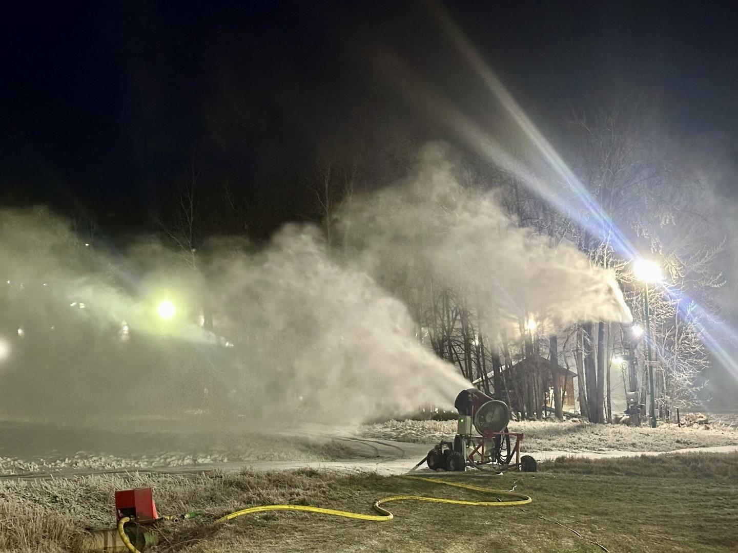 Snowmaking machines operating at night on a grassy field with lights illuminating the scene.