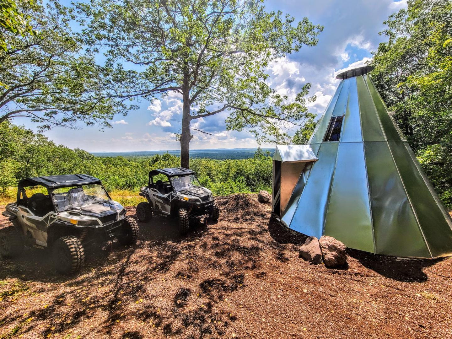 Two off-road vehicles beside a metal teepee in a wooded area under a clear sky.