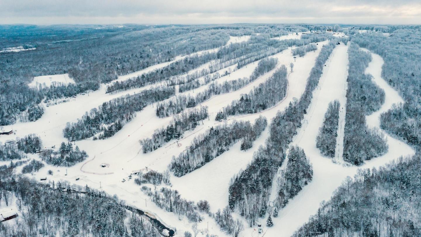 Snow-covered ski slopes surrounded by dense forest, under a cloudy sky.