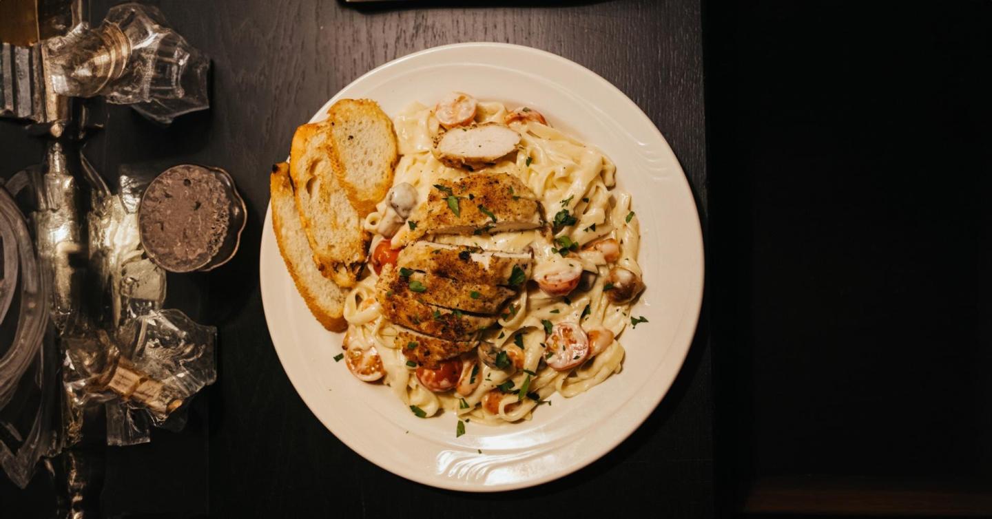 Grilled chicken pasta with toast, served on a white plate in dim lighting.